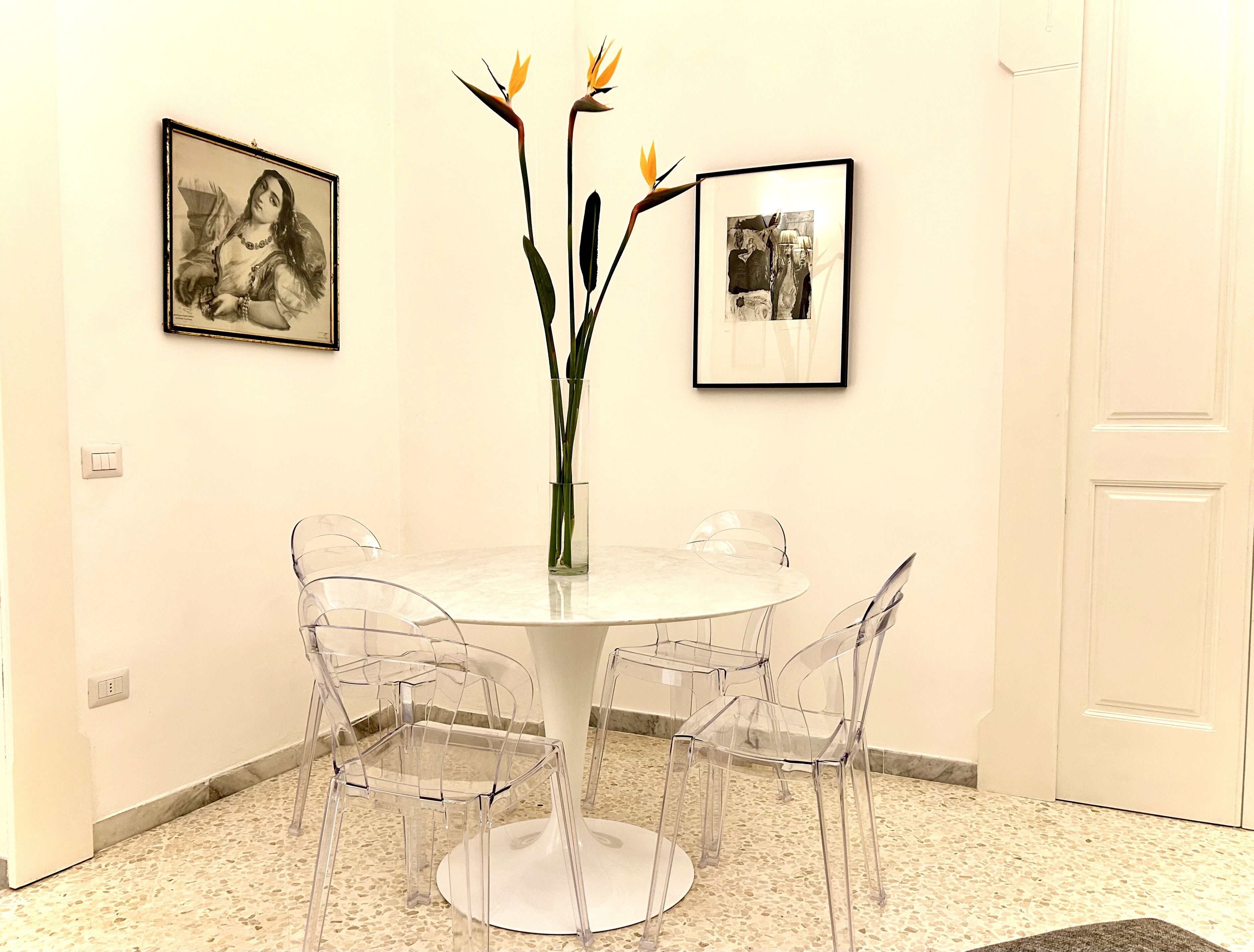 A round dining table with four clear chairs, a vase with flowers, and two framed pictures on the wall.