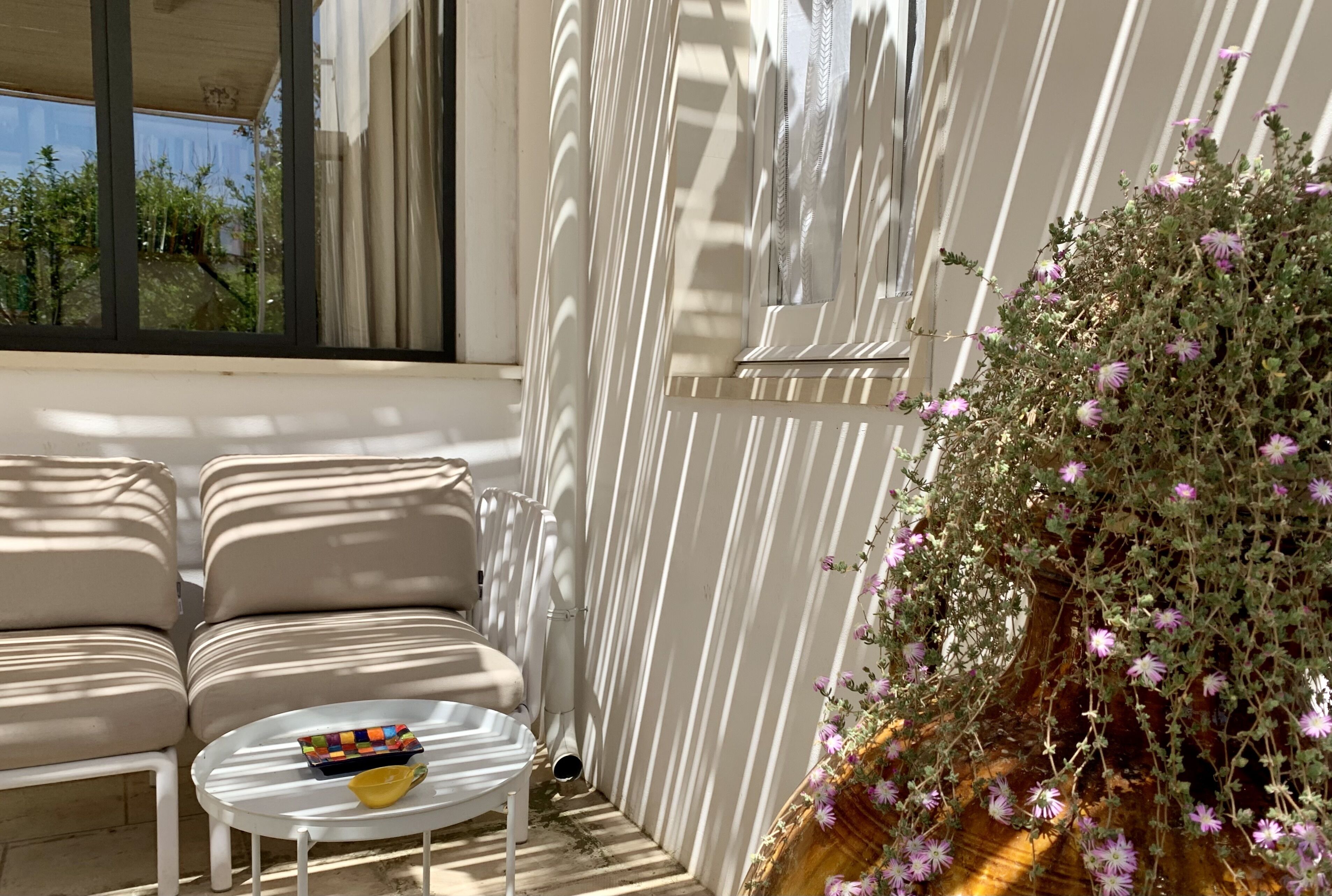Un área de estar al aire libre iluminada por el sol, con una mesa y sillas blancas, una maceta con flores moradas y vistas a follaje a través de una ventana.