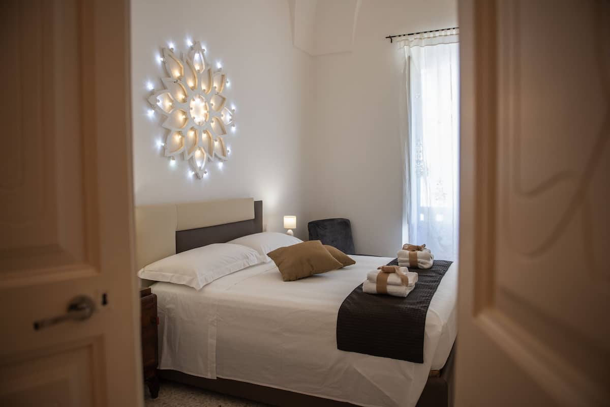Una habitación de hotel con una cama bien hecha, una lámpara de pared y una ventana con cortinas.