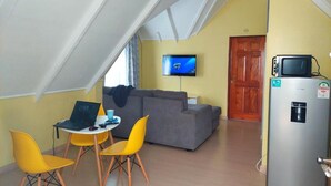 A compact living space with a lofted ceiling, a gray sofa, a small dining table with two yellow chairs, a laptop, a television, and a refrigerator. - Charming 1-bed Penthouse in Karen Nairobi (Nairobi)