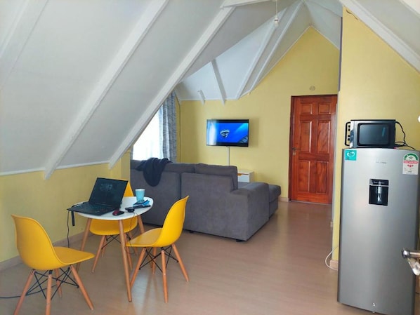 A compact living space with a lofted ceiling, a gray sofa, a small dining table with two yellow chairs, a laptop, a television, and a refrigerator.