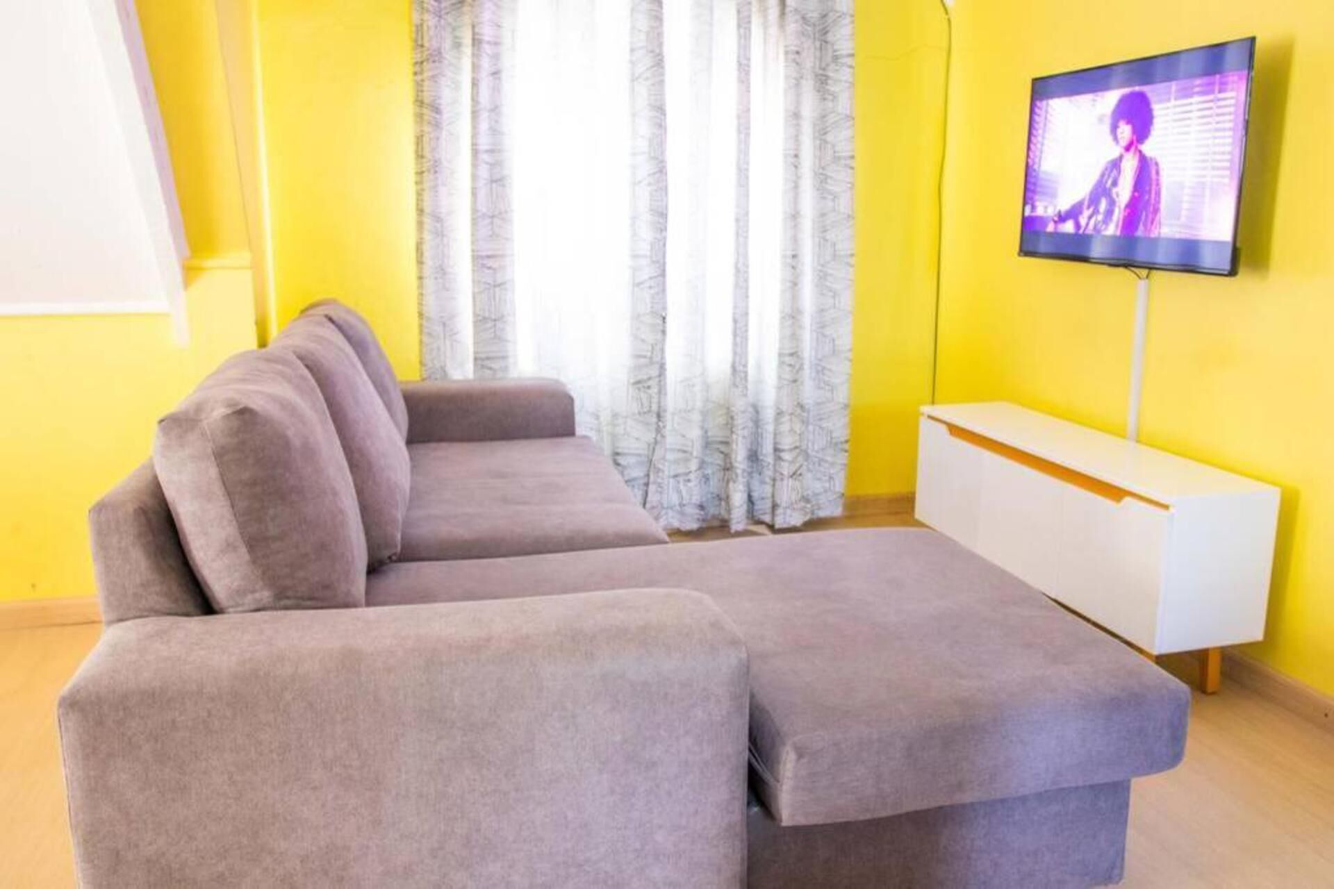 A living room with a grey sectional sofa, a white TV stand, and a yellow wall.