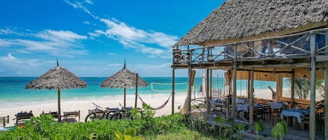 A beachside restaurant with thatched umbrellas, wooden tables, and a view of the ocean.