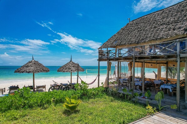 A beachside restaurant with thatched umbrellas, wooden tables, and a view of the ocean. - La Perla Beach Resort, Zanzibar - Your Beachfront Private Haven (Pwani Mchangani)