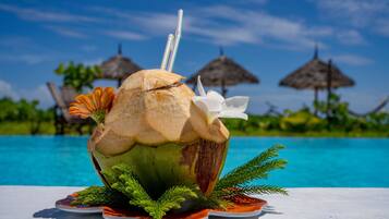 Bebida de coco con pajita y una flor, sobre un plato con follaje, cerca de una piscina con sombrillas al fondo.