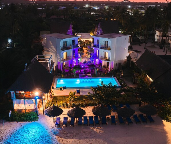 A beachfront property with a swimming pool, lounge chairs, and a well-lit building. - La Perla Beach Resort, Zanzibar - Your Beachfront Private Haven (Pwani Mchangani)