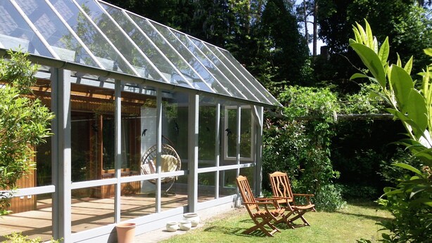 A conservatory with a glass roof, wooden chairs, and a garden view.