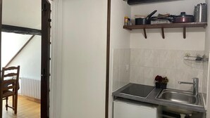 A compact kitchen with a built-in stove, a sink, and a small refrigerator. There is a wooden chair in the adjacent dining area, and a shelf above the sink holding kitchen utensils. - Studio Mezzanine 4 Personnes (Hargeville)