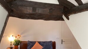 A bedroom with a sloped ceiling, wooden beams, a bed with patterned bedding, a nightstand with a lamp and flowers, and a door. - Studio Mezzanine 4 Personnes (Hargeville)