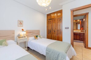 A hotel room with two beds, a nightstand with a lamp, a wooden wardrobe, and a bathroom visible through an open door.