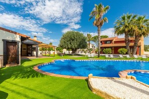 Una piscina con un patio alrededor, una zona de estar techada y un edificio de techo rojo.