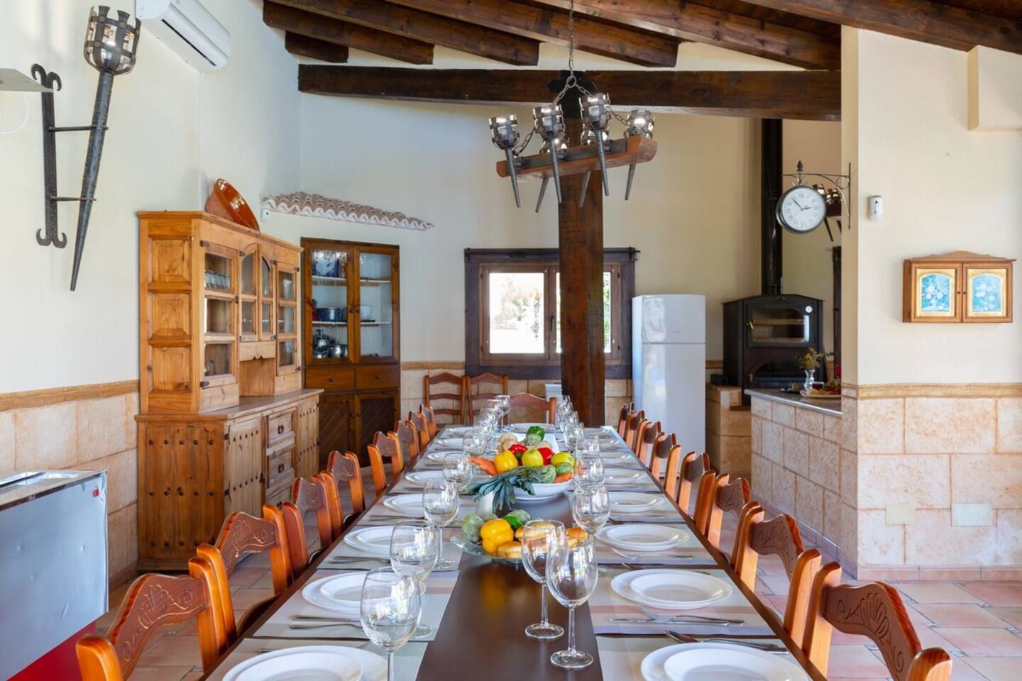 Un comedor con una larga mesa dispuesta para una comida, armarios de madera, una araña y un reloj en la pared.