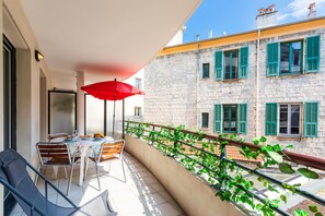 A balcony with a table set for dining, a red umbrella, and a view of a stone building with green shutters.