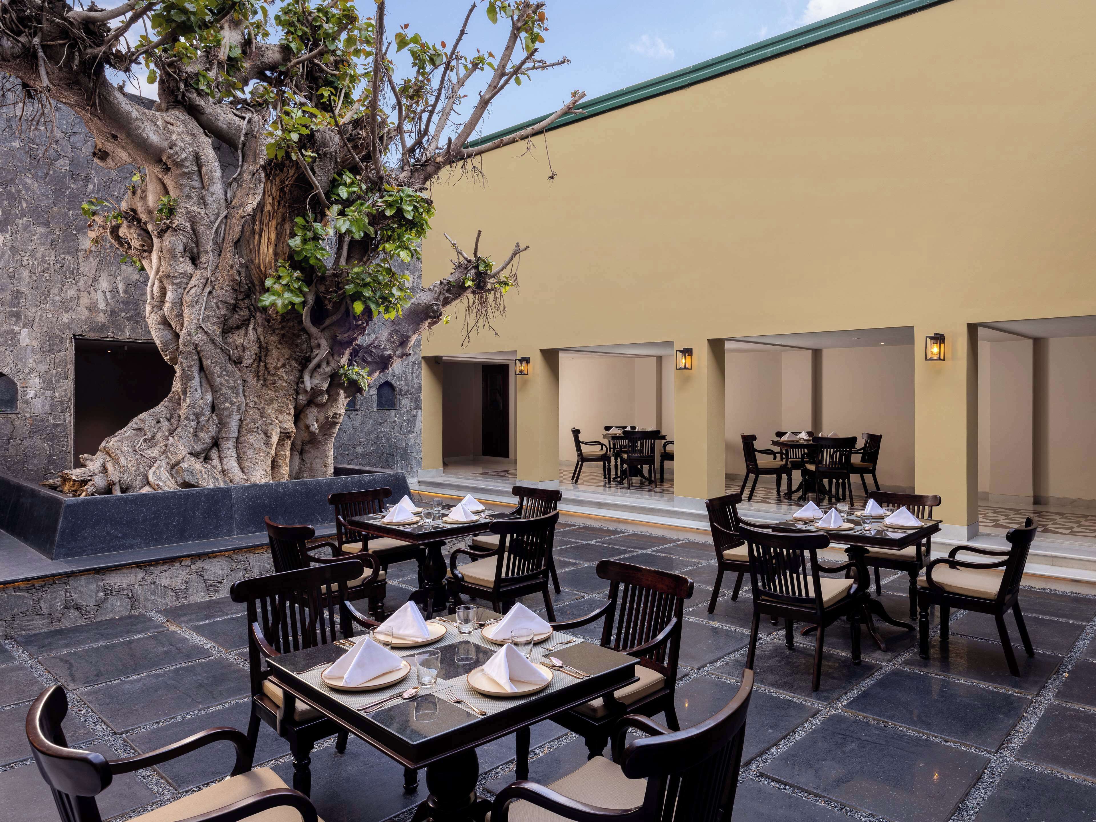 Un espacio al aire libre con mesas dispuestas para cuatro personas, rodeado de muros de piedra y un gran árbol.
