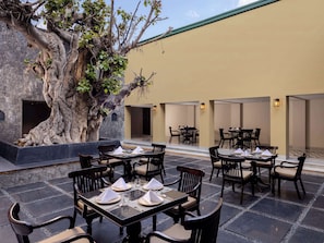 Un espacio al aire libre con mesas dispuestas para cuatro personas, rodeado de muros de piedra y un gran árbol.