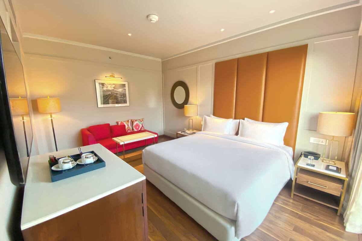 Ein Hotelzimmer mit einem großen Bett, einem roten Sofa, einem Schreibtisch mit einem Tablett Gläser und einem an der Wand hängenden Bild.