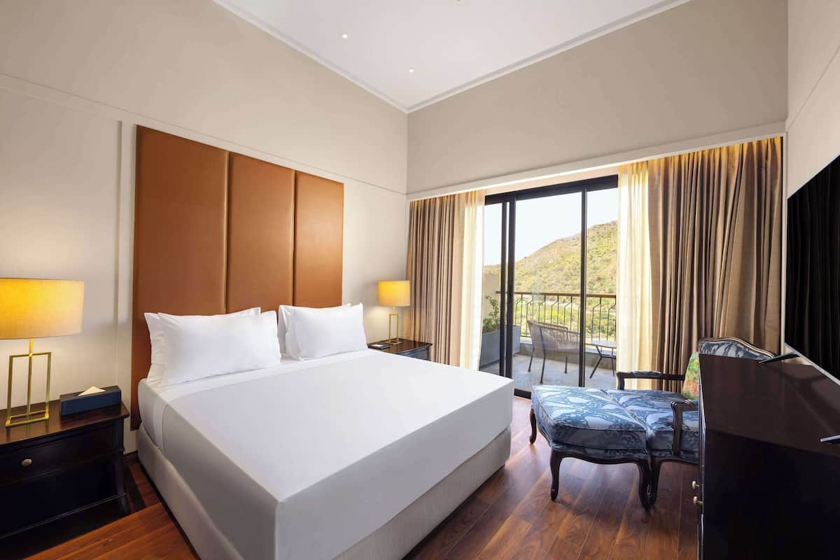 Ein Hotelzimmer mit einem großen Bett, zwei Nachttischen mit Lampen, einem Schreibtisch mit Stuhl und einem Balkon mit Vorhängen.