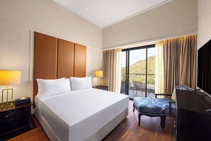 Habitación de hotel con una cama grande, dos mesitas de noche con lámparas, un escritorio con silla y un balcón con cortinas.