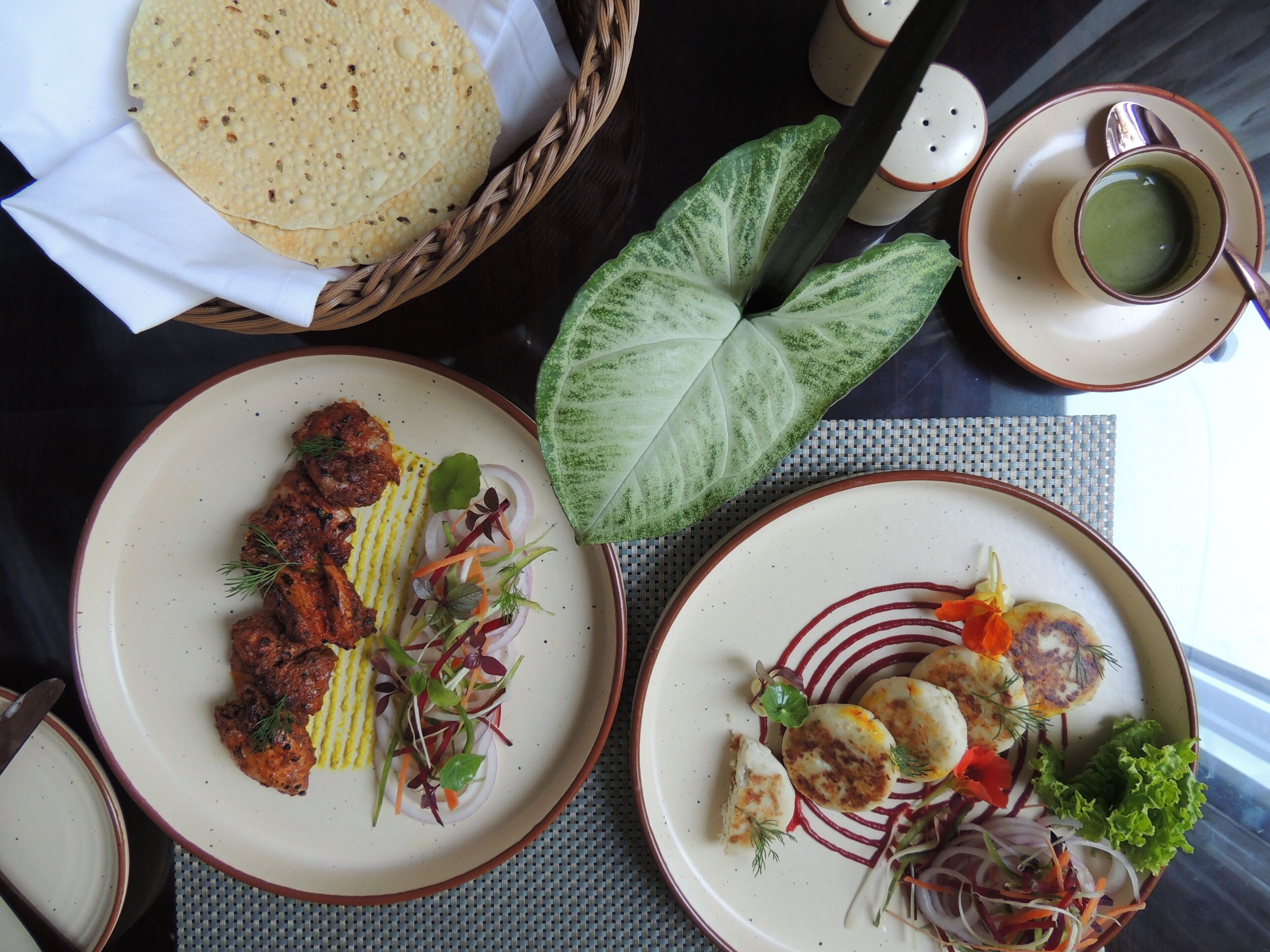 Una mesa de comedor con platos de comida, una cesta de pan plano y una taza de té verde.