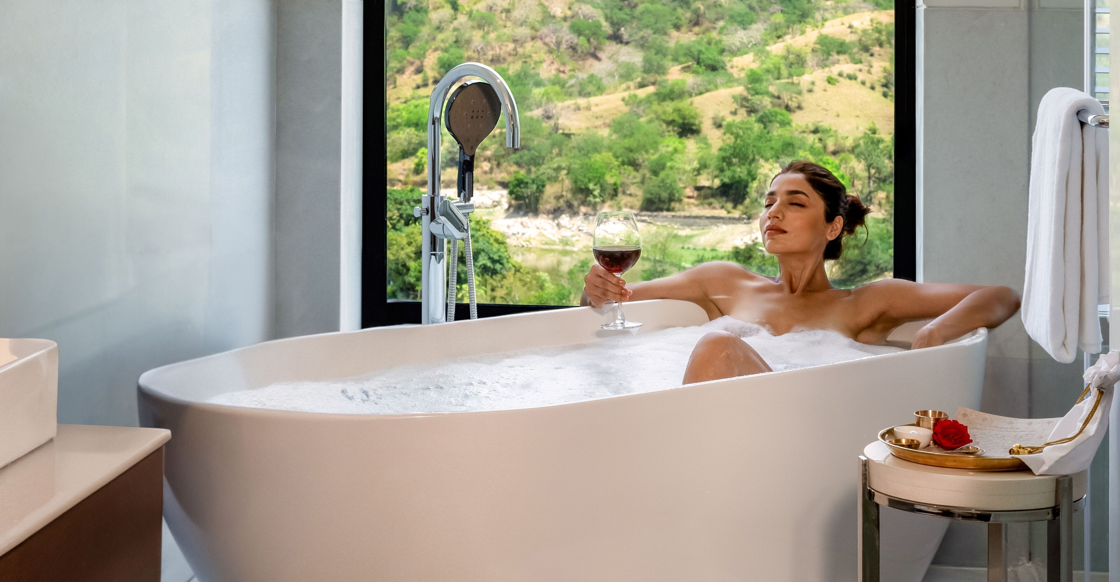 A person relaxing in a bathtub with a view of a green landscape through a window.
