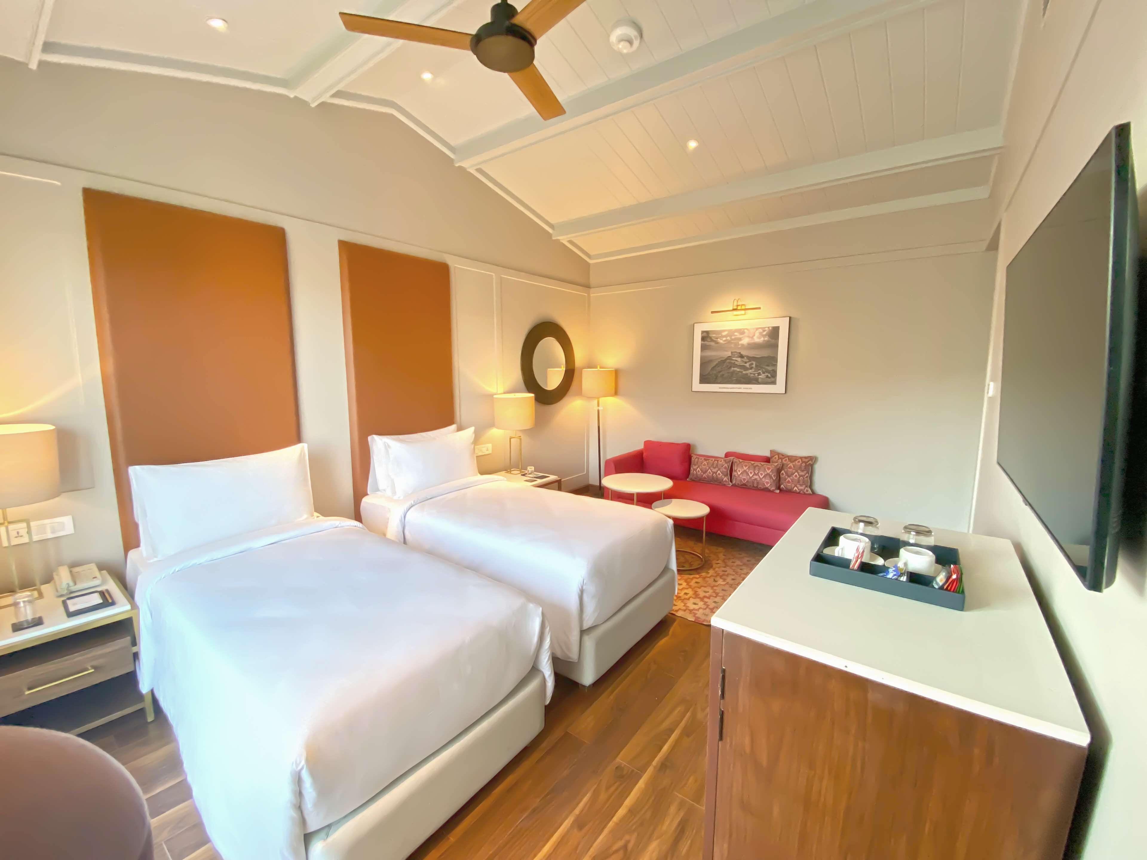 A hotel room with two beds, a red sofa, a desk with a TV, and a ceiling fan.