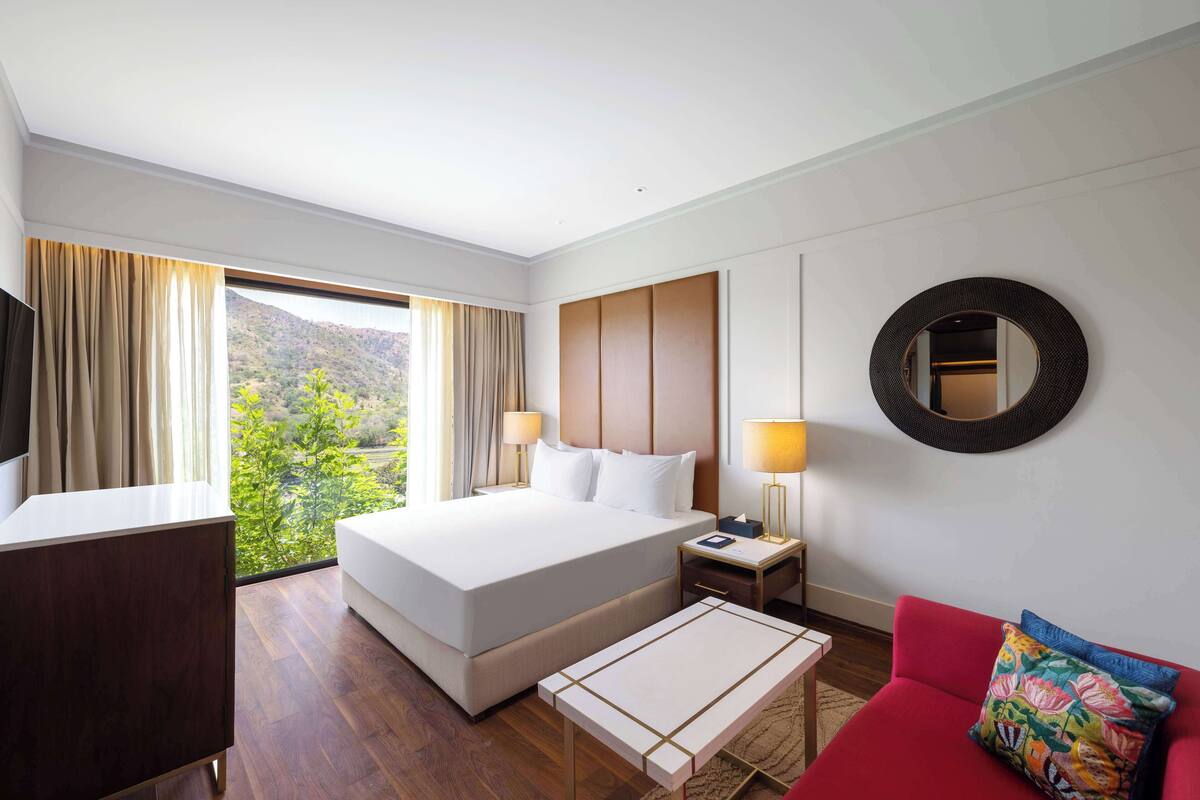 Ein Hotelzimmer mit einem großen Bett, einem Schreibtisch, einem roten Sofa und Blick auf Grün hinter dem Fenster.
