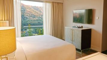 Habitación de hotel con una cama grande, un televisor en la pared, un escritorio con lámpara y vistas a una montaña y un lago.