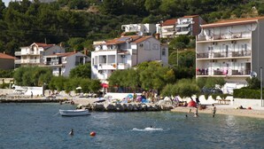 Une ville côtière avec des bâtiments le long du front de mer, notamment un hôtel doté de balcons et une plage animée où l’on peut voir des gens et des bateaux.