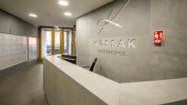 Een receptiebalie met een bord waarop 'KOSSAK RESIDENCE' staat en een brandmelder aan de muur.