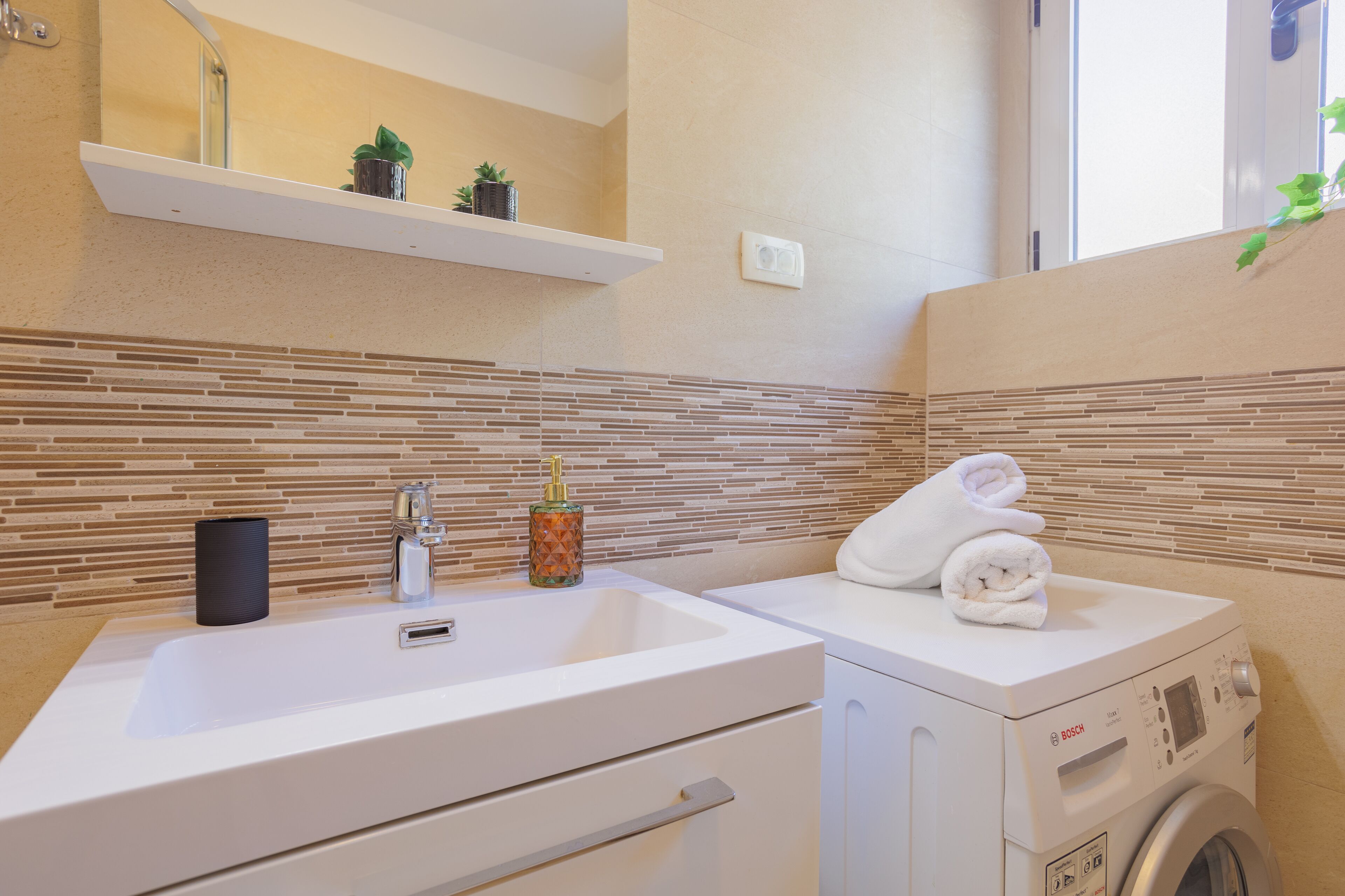 A modern bathroom with a white sink, a built-in washing machine, and a tiled backsplash.