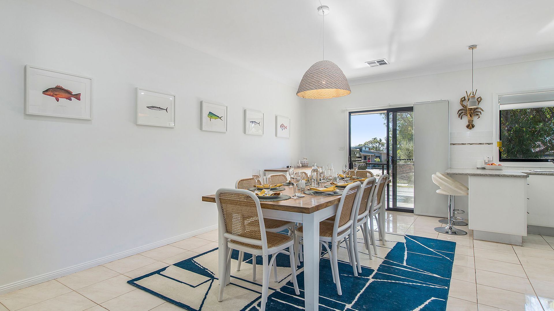 Un coin repas avec une grande table dressée pour six personnes, entourée de chaises en osier, et un tapis à motifs bleus sur le sol.