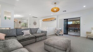 Een moderne woonkamer met een grijze modulaire bank, een beige fauteuil en een klein voetenbankje. Er hangt een houten surfboard aan de muur, een ronde wanddecoratie en een plafondlamp.