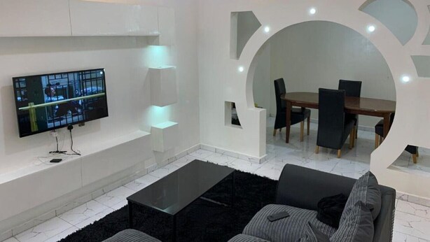 A modern living room with a flat-screen TV, a coffee table, and a dining area with chairs and a table. - Matano Guest House (Brikama)
