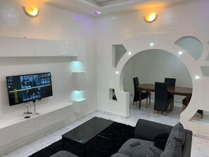 A modern living room with a flat-screen TV, a coffee table, and a dining area with chairs and a table.