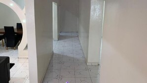A hallway with white marble flooring, a dining area with a table and chairs, and a door at the end. - Matano Guest House (Brikama)