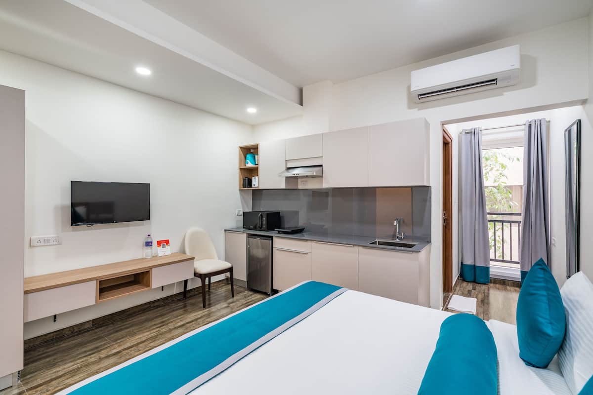 A modern hotel room with a bed, a desk, a television, a kitchenette, and a balcony with curtains.