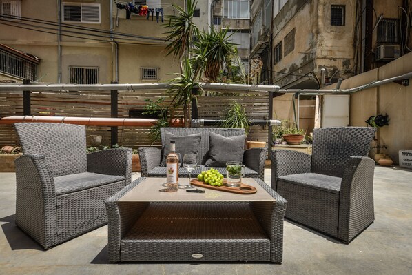 An outdoor seating area with wicker furniture, a table with a bottle, glasses, and fruit. - Unique and Spacious 2BR Apt with Terrace (Tel Aviv)