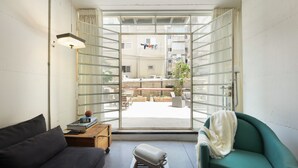 Una sala de estar moderna con un sofá, un sillón verde, una mesita baja con libros y una pelota, y una ventana con vista al balcón exterior.