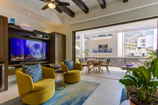 Ein modernes Wohnzimmer mit Flachbildfernseher, gelben Sesseln, einem Essbereich und einem Balkon mit Blick auf Gebäude und Grünflächen.