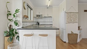 A modern kitchen with white cabinets, a microwave, and a refrigerator. There are two stools at the counter and a plant on the left.