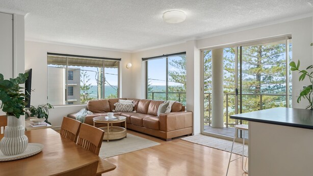 A modern living room with a leather sofa, wooden dining table, and large windows offering a view of the sea.