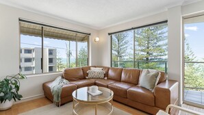 A modern living room with a brown leather sectional sofa, a glass coffee table, and large windows offering a view of buildings and trees.