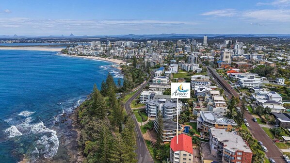 A coastal cityscape with a mix of residential and commercial buildings, a prominent hotel with a red roof, and a clear view of the ocean. - Minya Court Unit 4 Kings Beach QLD (Kings Beach)