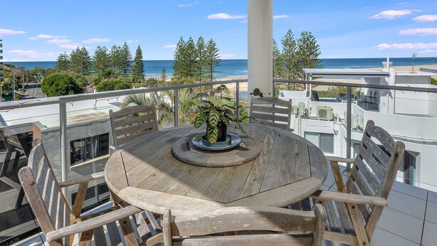 Une table ronde en bois, sur laquelle est posée une plante en pot, se trouve sur un balcon donnant sur une plage.