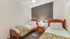 Une chambre avec deux lits en bois, une table de chevet, une fenêtre avec des stores et un ventilateur de plafond.