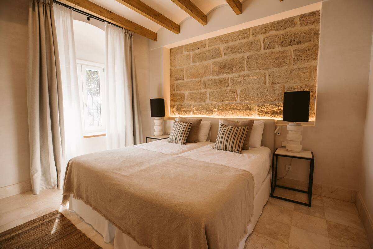 Un dormitorio con una cama grande, dos mesitas de noche con lámparas, una pared de piedra y una ventana con cortinas.
