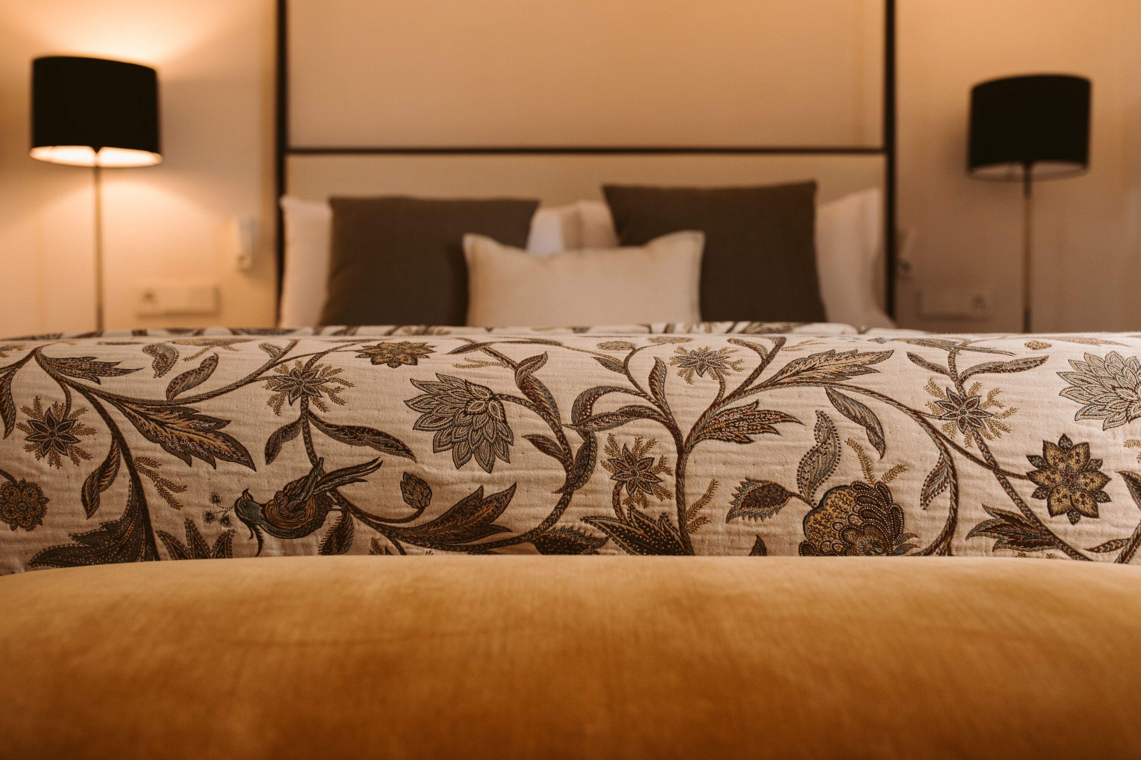 Una cama bien hecha con una colcha estampada de flores, dos lámparas de noche y un cabecero.