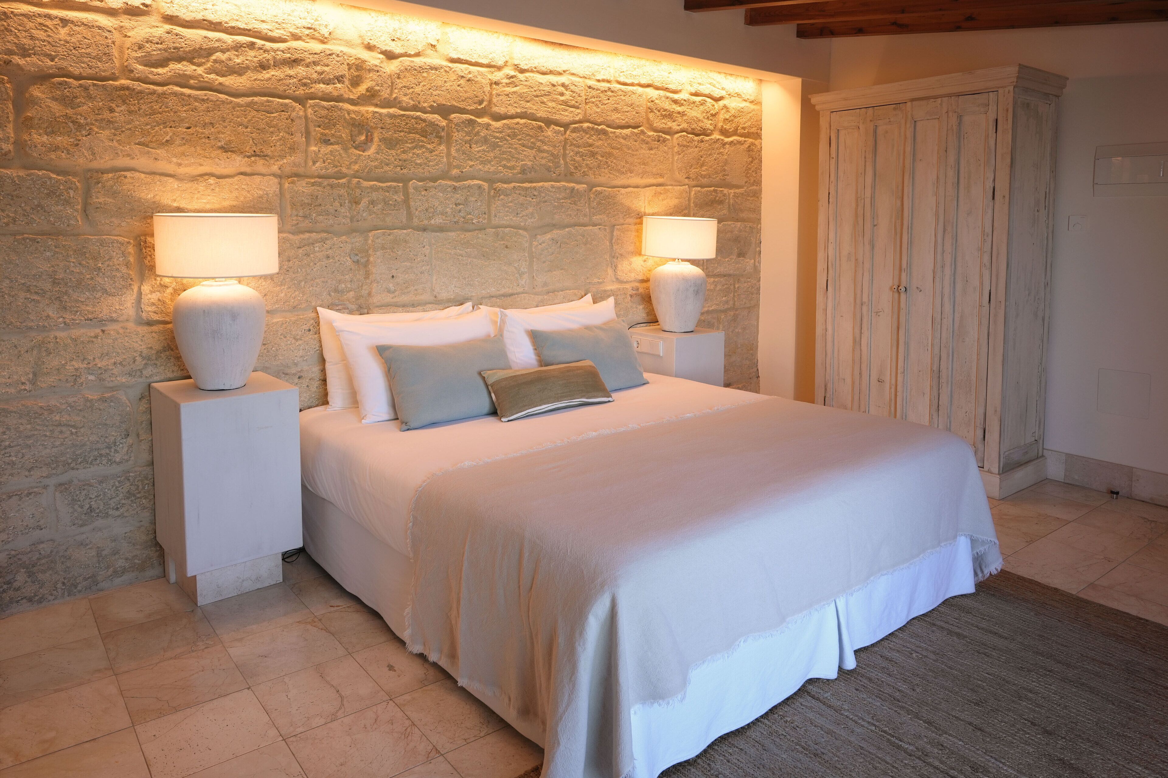 A bedroom with a stone wall, a bed with white bedding, two bedside lamps, and a wooden wardrobe.