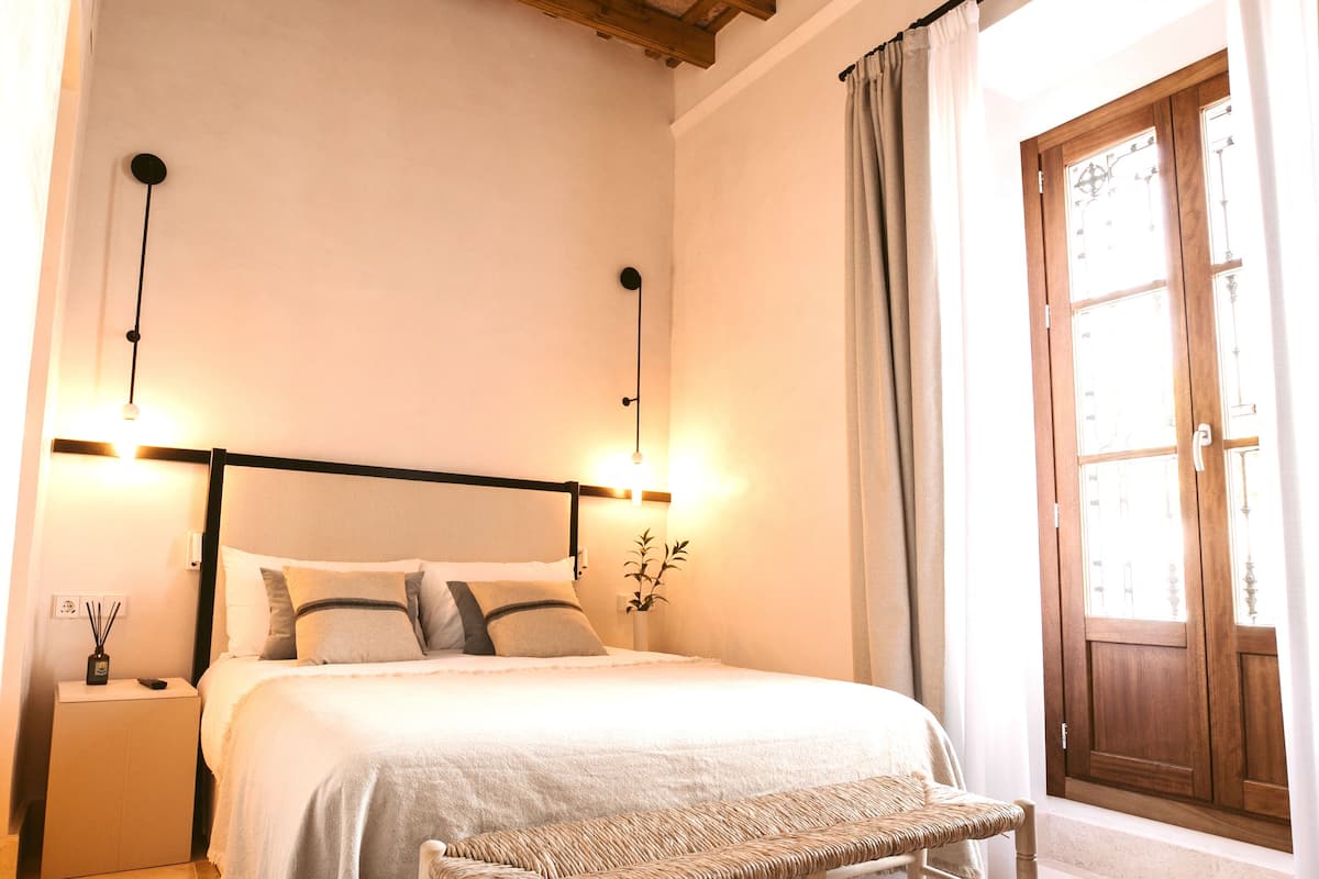 Un dormitorio con techo de madera, cama con ropa de cama blanca, mesita de noche con lámpara y ventana con cortinas.