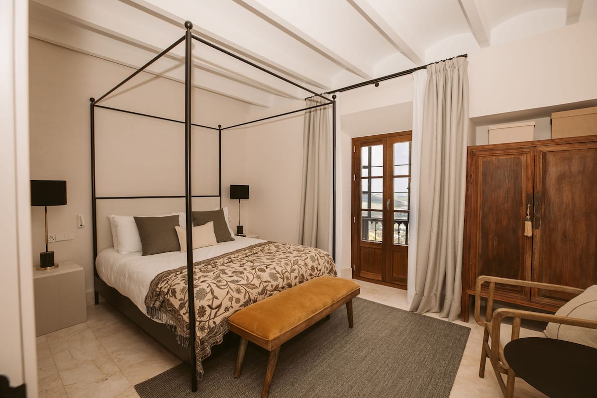 Una cama con dosel, un banco, un armario de madera y vistas al exterior a través de una puerta.